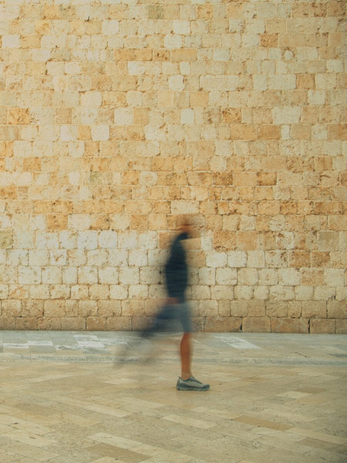 A blurred person walking past a textured stone wall on a paved plaza.